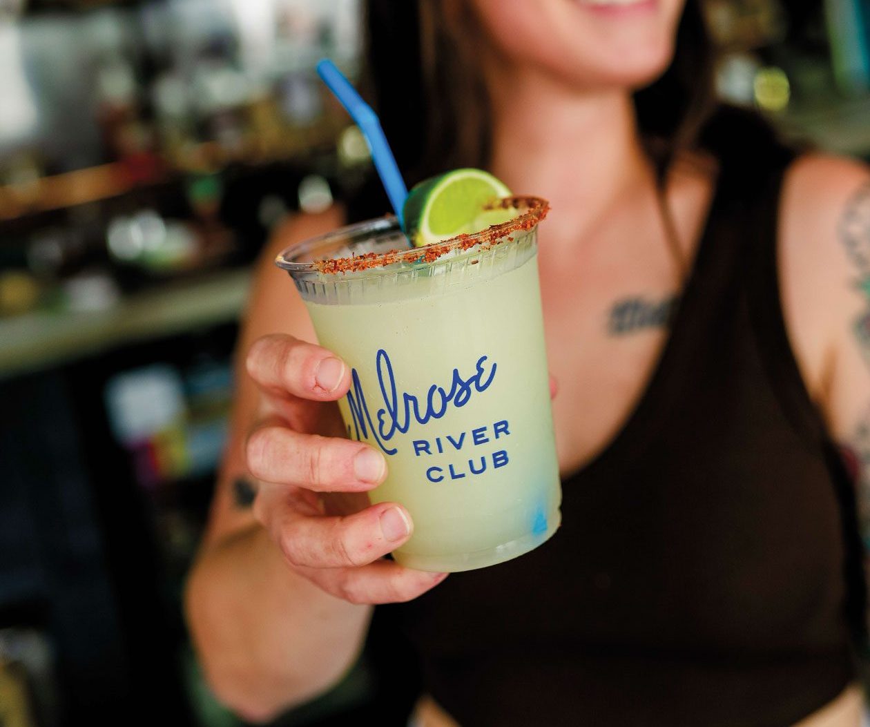 A woman holds a margarita up to the camera and the cup reads "Melrose River Club"